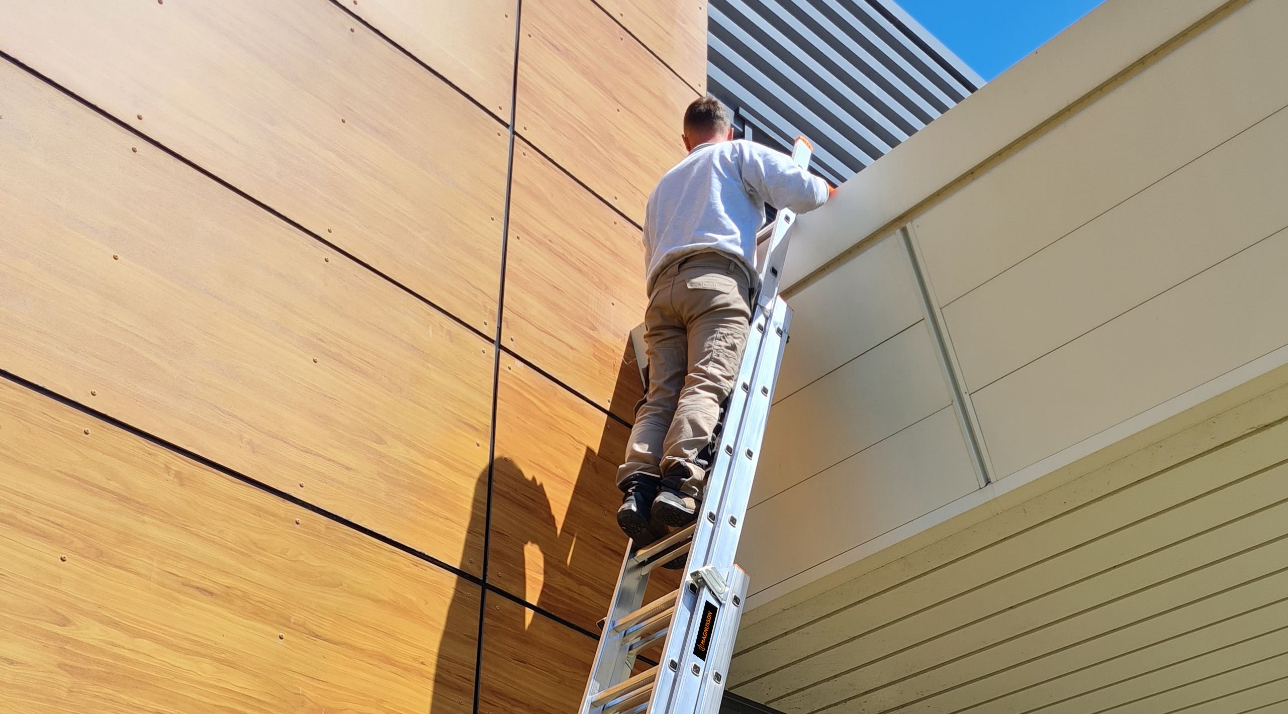 Man climbing a silver ladder against a modern building with wooden paneling.