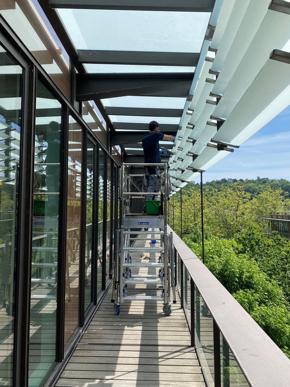 Worker on scaffolding cleans glass canopy outside modern building with trees.