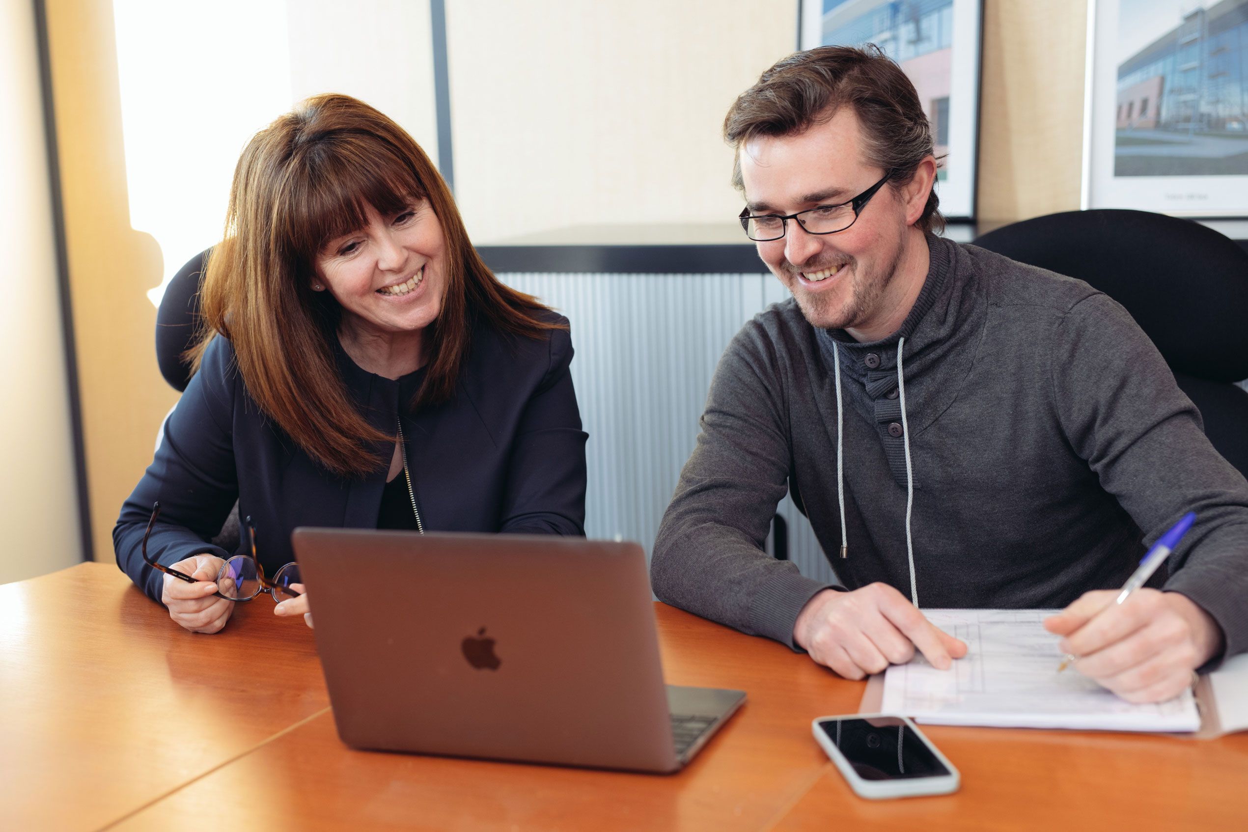 Two smiling colleagues collaborate on a laptop in a modern office meeting.