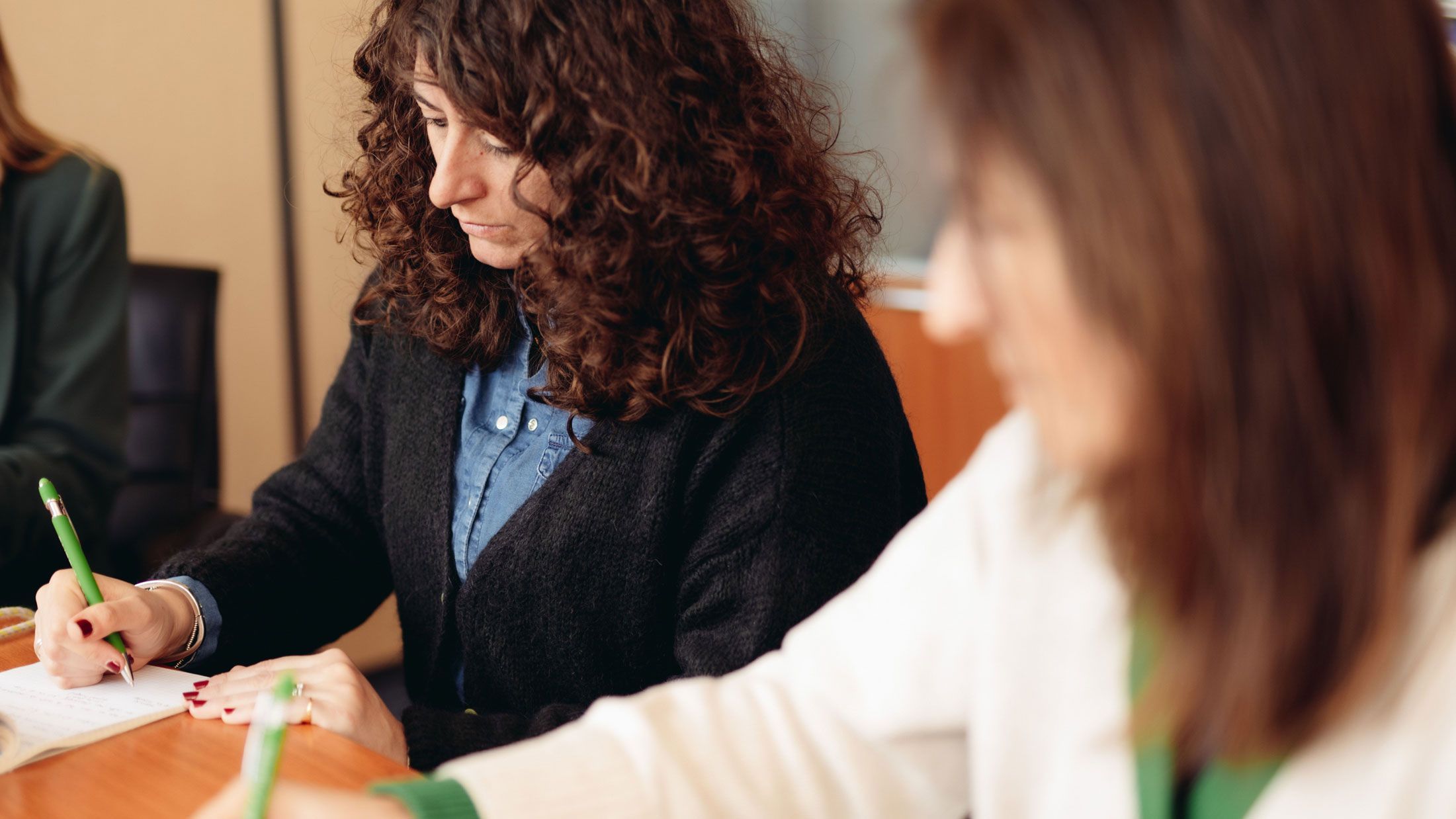 A woman with dark curly hair writes notes with a green pen at a table.