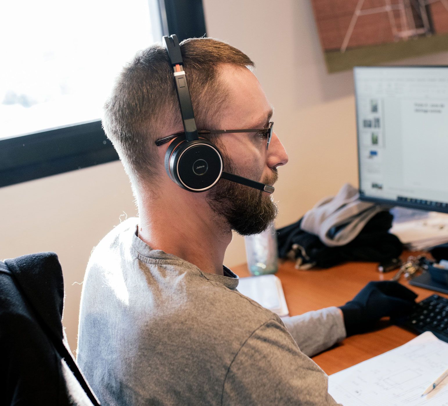 Bearded man wearing glasses and a headset works at a computer in an office.