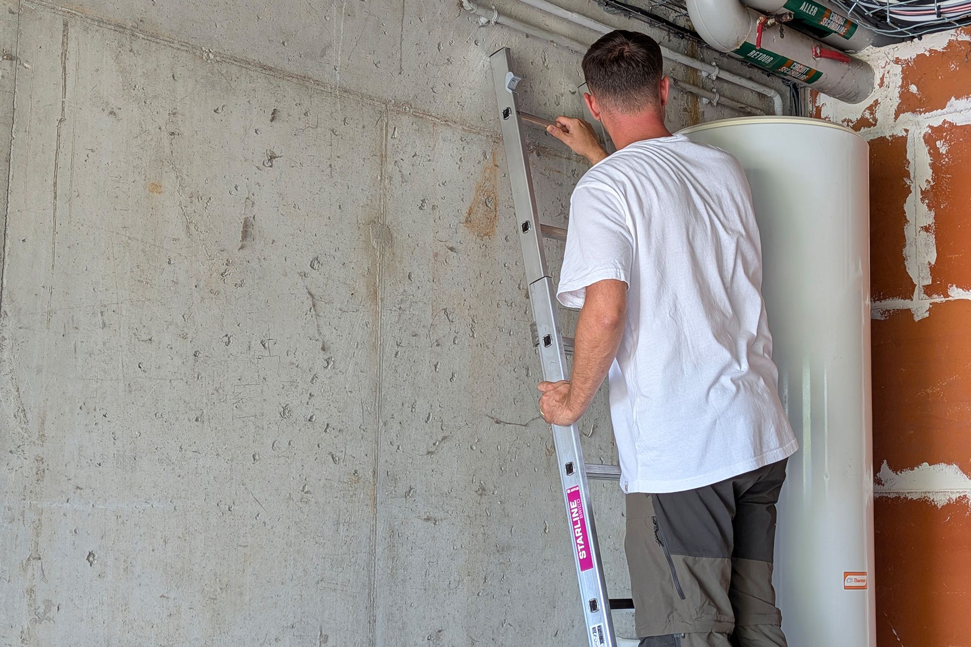 Man on a ladder working near pipes against a concrete wall.