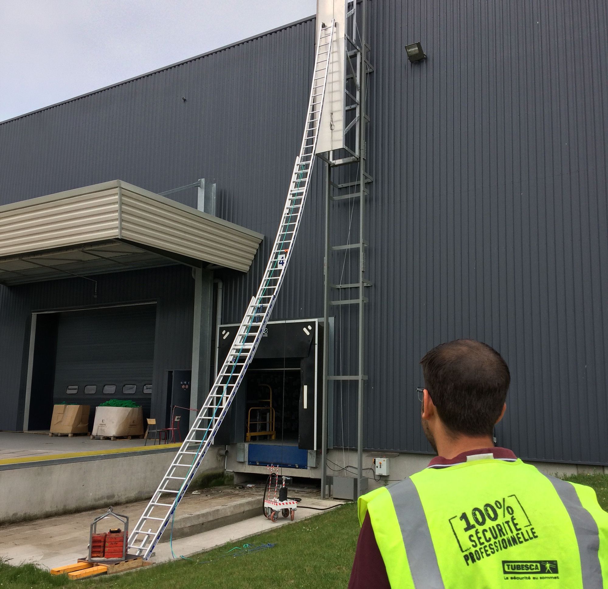 A man watches a very long, multi-section ladder extending up a grey industrial building.