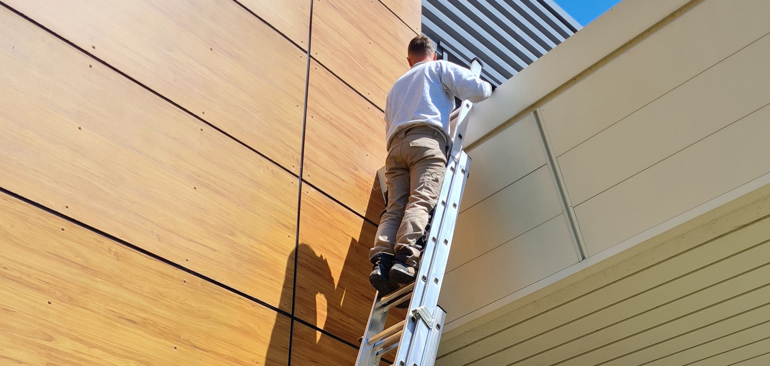 Man on a ladder working on the exterior of a modern building with wooden panels.