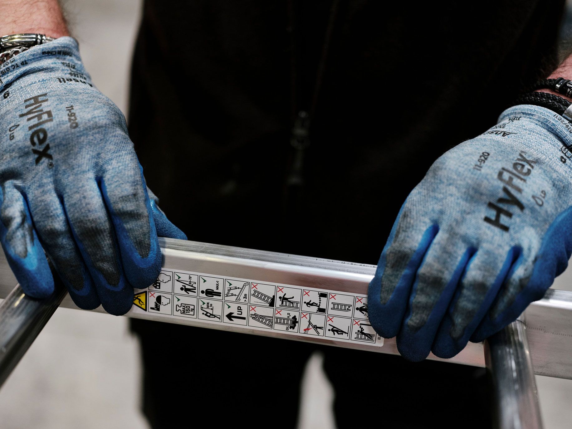 Person wearing blue HyFlex gloves firmly grips a silver metal ladder with safety labels.
