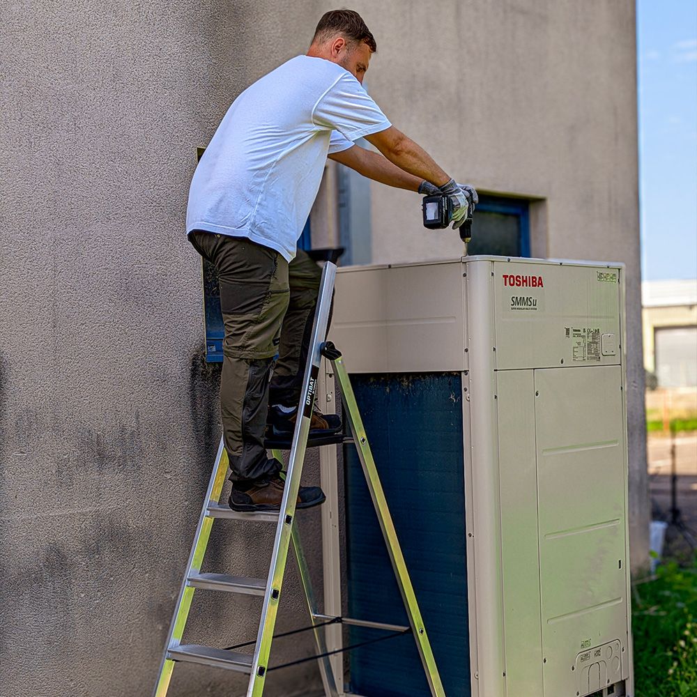 Man on ladder servicing an outdoor Toshiba HVAC unit against a building wall.