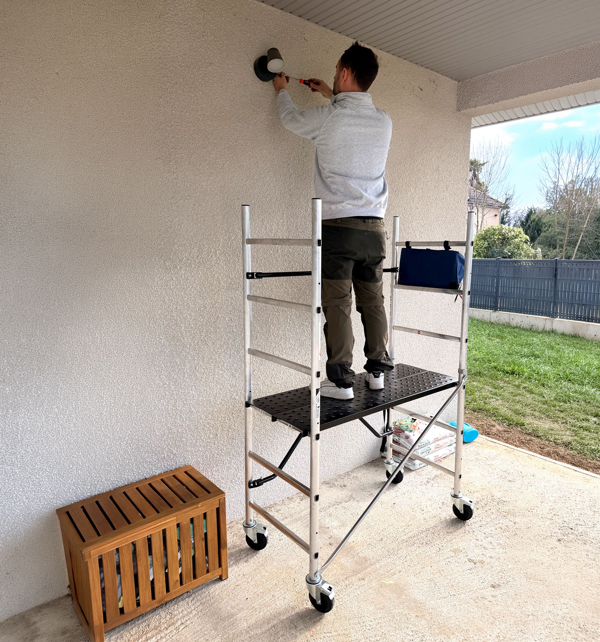A man on a mobile scaffold is drilling or sanding a white exterior wall.