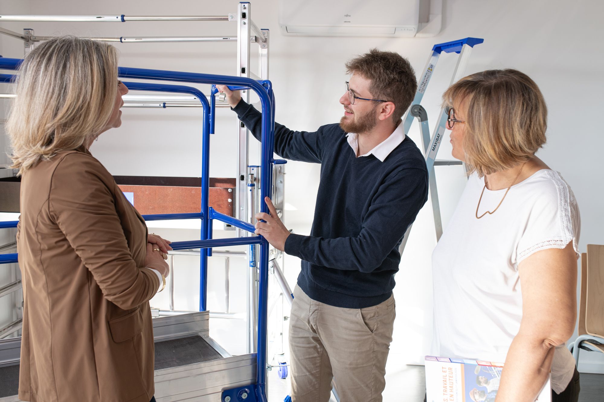 A man wearing glasses explains a blue metal scaffolding system to two attentive women.