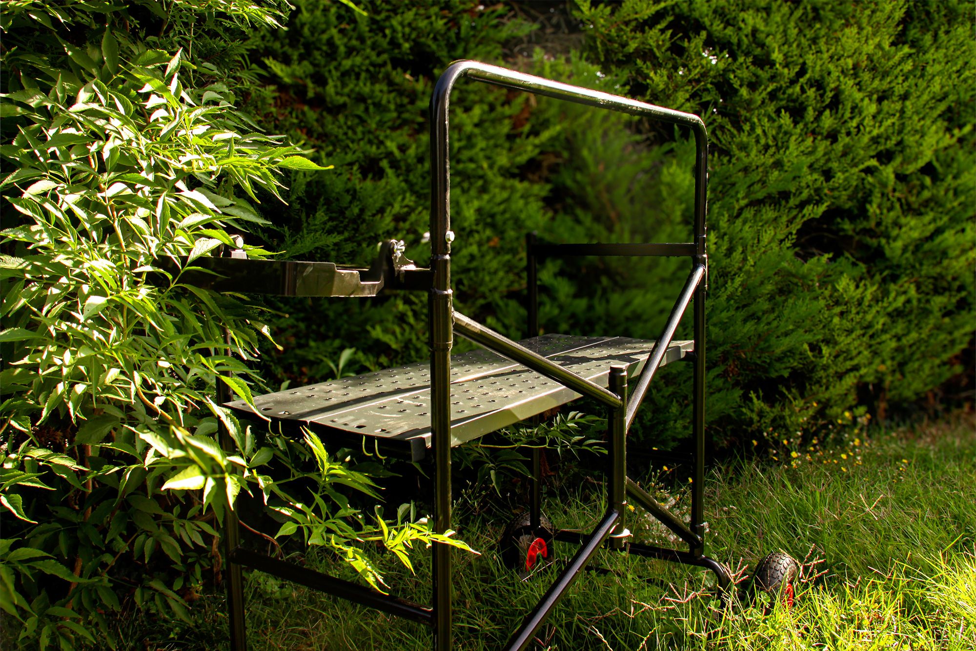 A portable black metal work platform stands in a sunlit garden among green foliage.