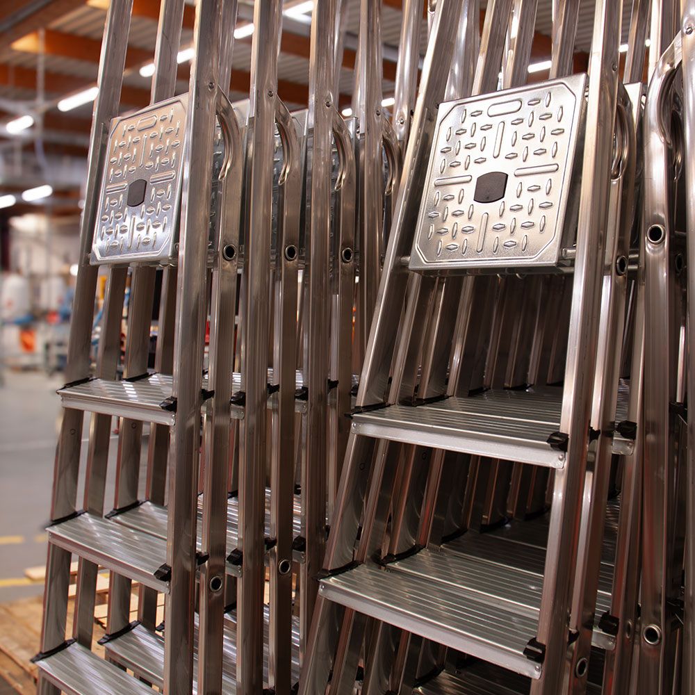 Multiple silver aluminum stepladders are neatly stacked and stored in a warehouse.