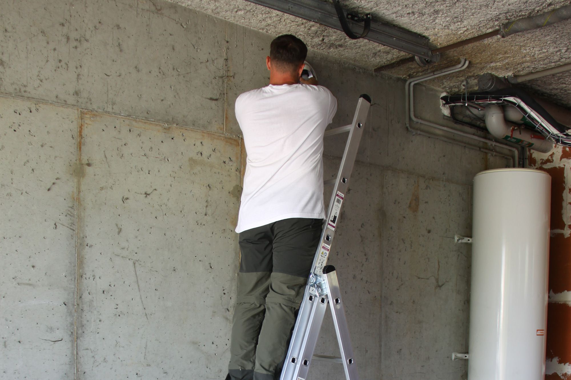 Man on a ladder installing pipes or wiring against a concrete wall in a utility room.
