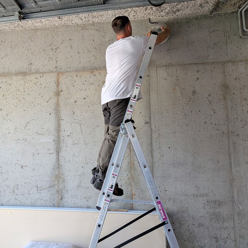 Man on a silver ladder reaching up to a concrete ceiling, performing maintenance work.